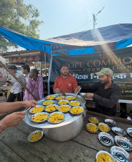 Hands reach in as volunteers serve plates of yellow rice from a large steel pot at a street stall, with a green Hope Commoners Foundation banner behind.