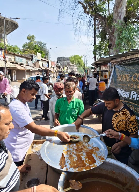 A man in a white shirt ladles food onto a plate across a steel pan while others look on, on a busy street in daylight.