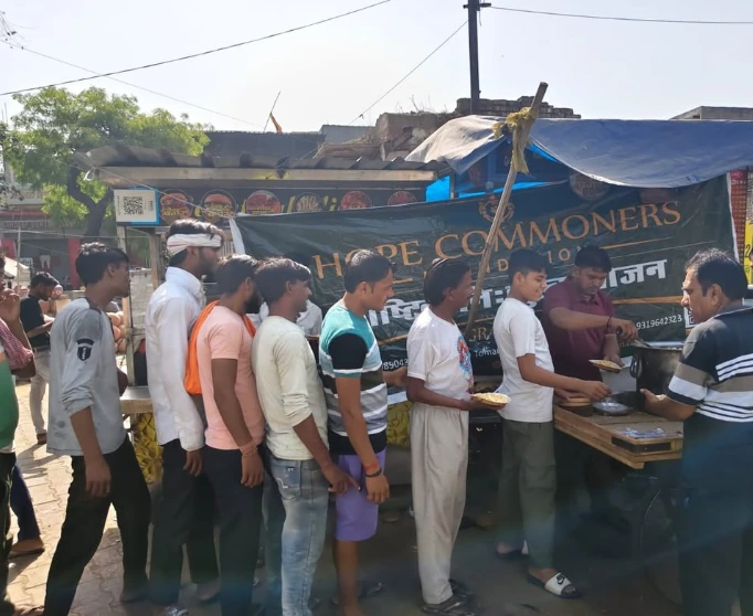 A line of people stands in a street beside a roadside stall with a green Hope Commoners Foundation banner.