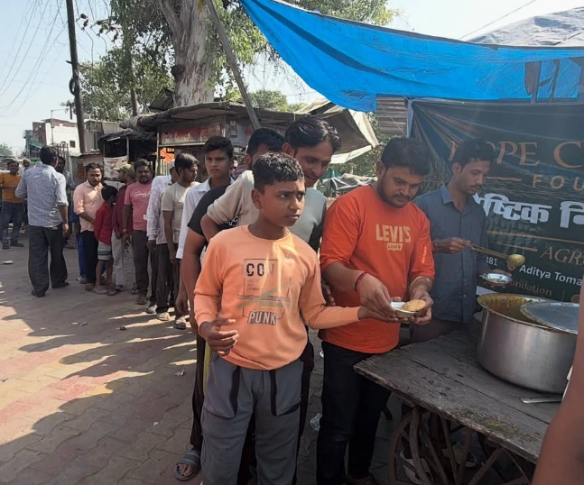 A boy in an orange top holds a plate at the serving counter while a queue forms behind him beneath a blue tarpaulin.