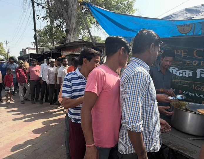 Men in checked and striped shirts wait at a serving counter beside a large pot, with a queue behind them.