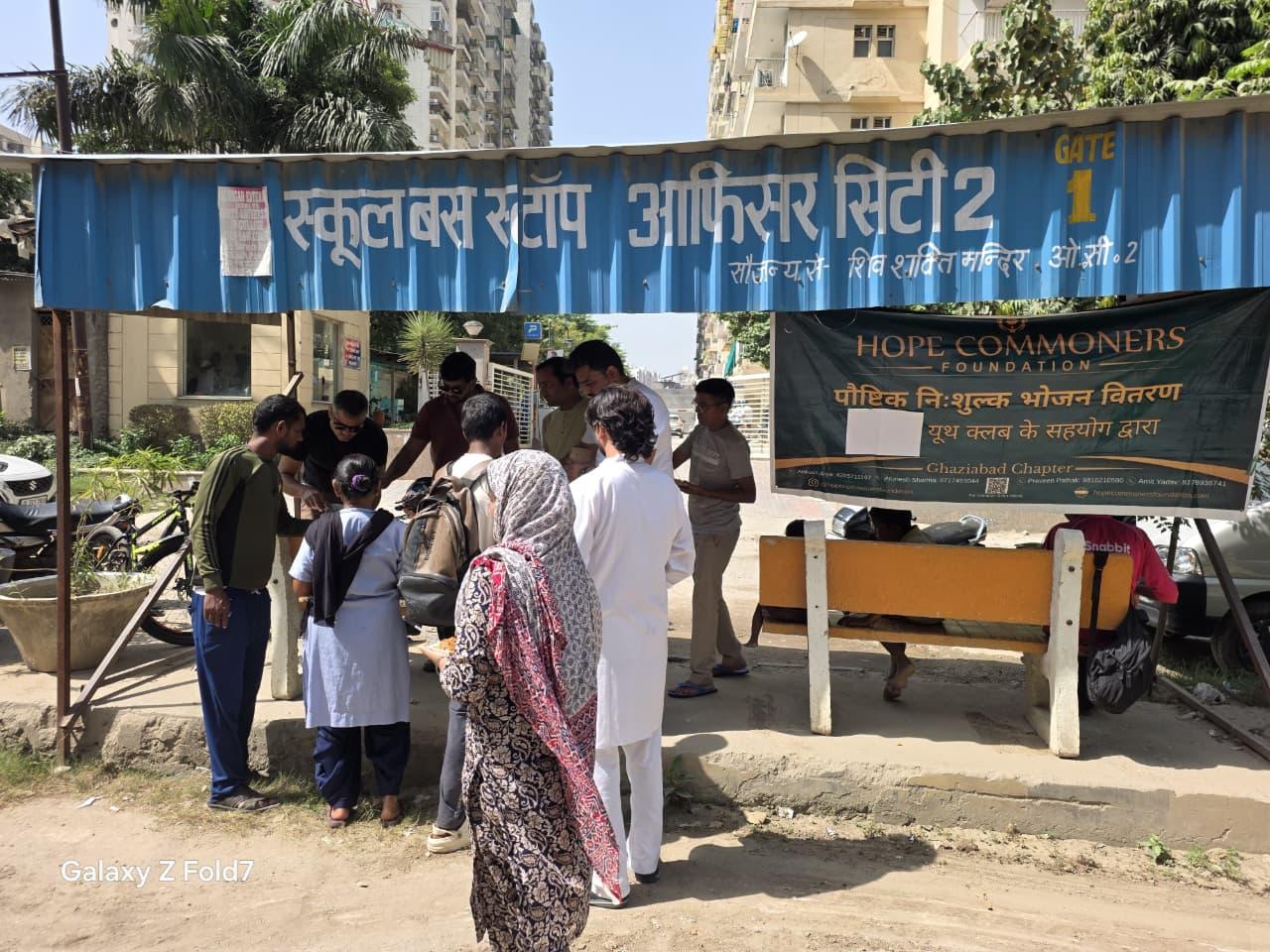 A shelter under a blue bus-stop sign with a green Hope Commoners Foundation banner reading Ghaziabad Chapter, with people gathered below.