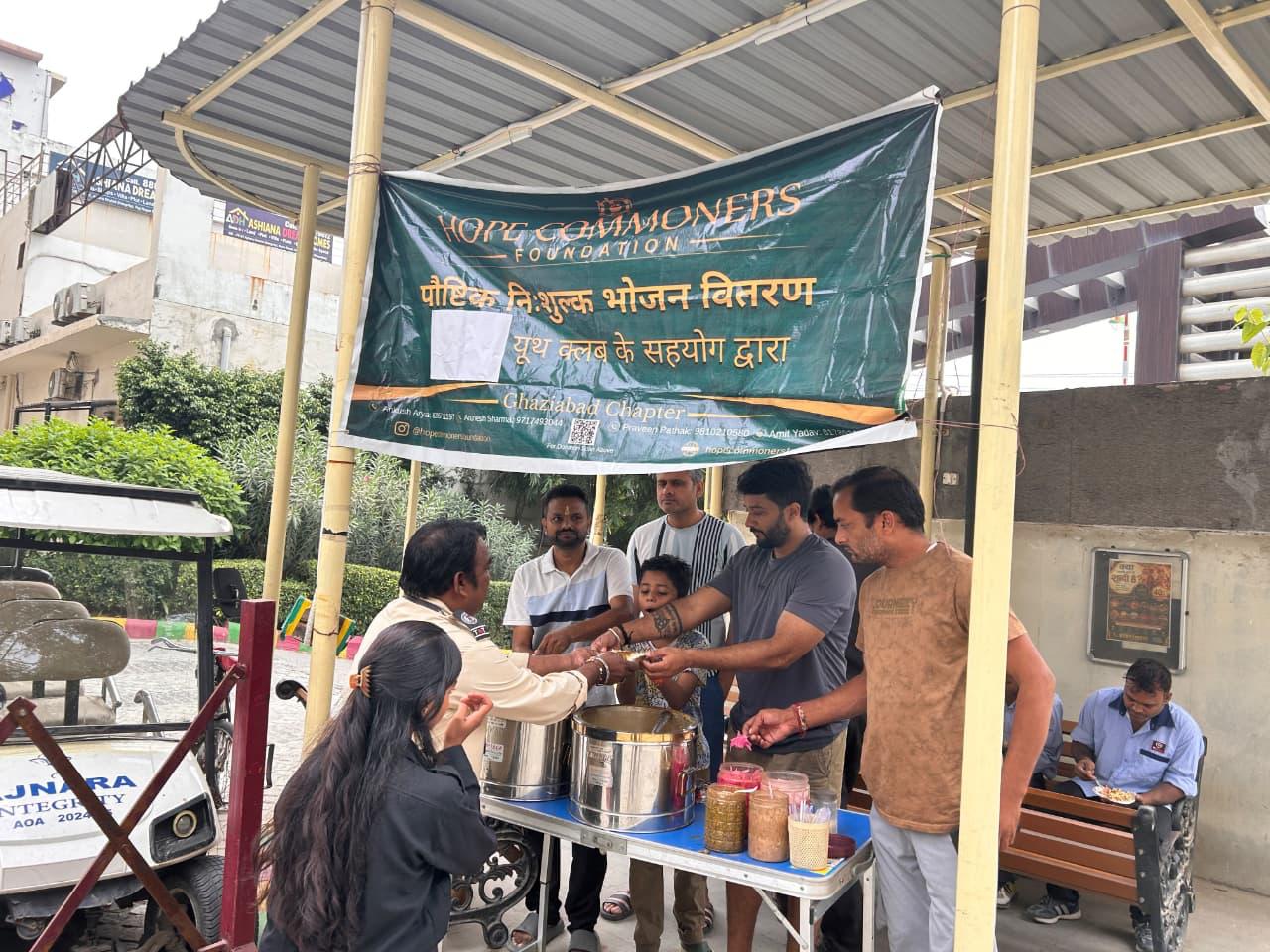 People serve and receive meals under a shelter roof and a Hope Commoners Foundation banner, with trees and apartment towers beyond.