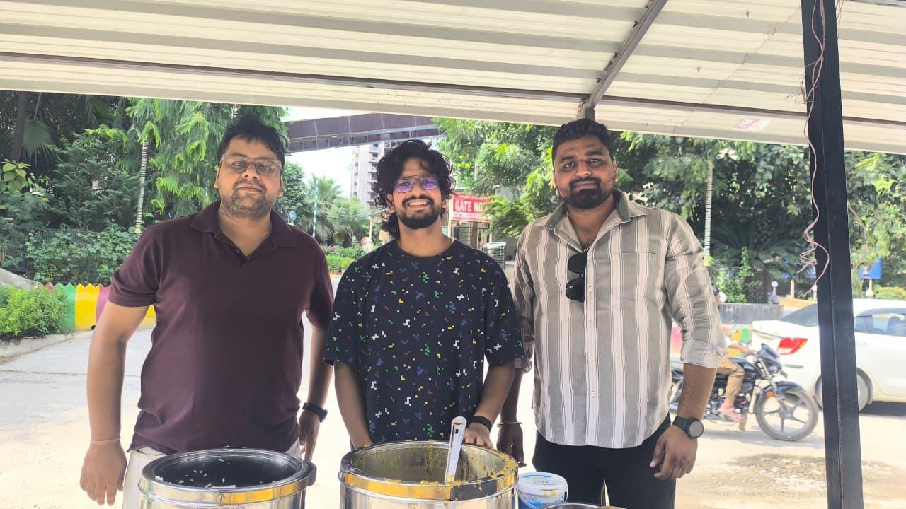 Three men smile at the camera behind two large steel serving pots under a shelter roof.