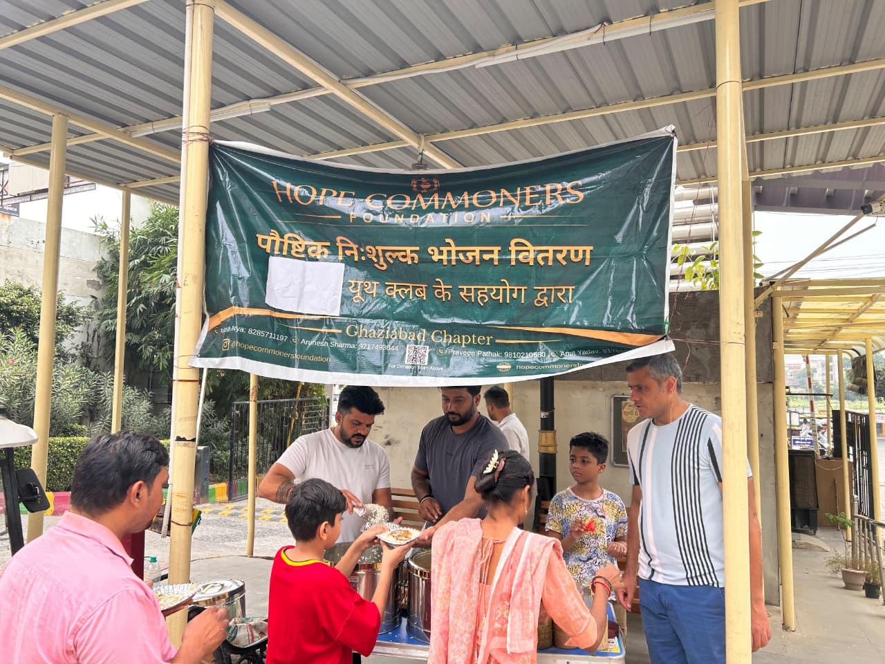 A woman in an orange kurta, a boy and other people receive plates from men serving at a counter under a green banner.