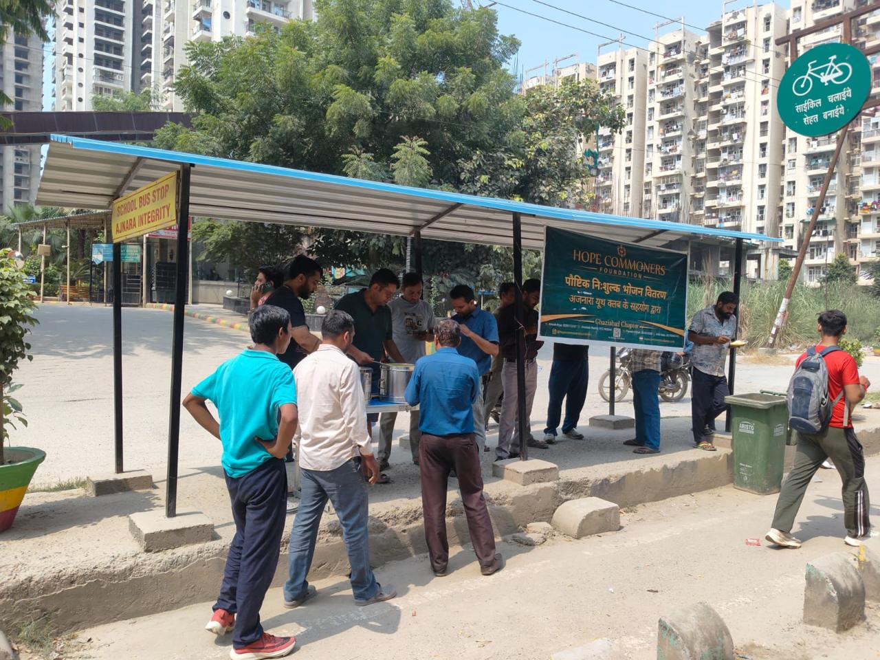 A shelter with a green banner and a group of people gathered at its counter, with apartment buildings and trees behind.