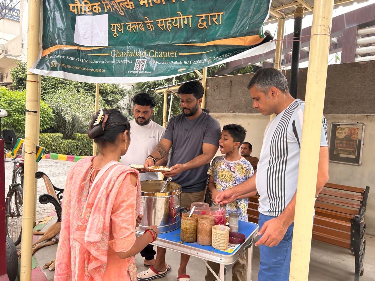 A woman in a peach kurta and a boy holding a plate stand at a counter with jars and steel containers, beside men serving.