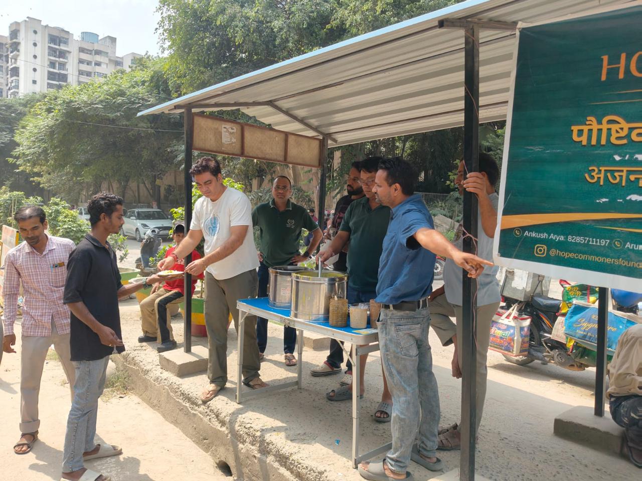 A group of people serve and receive food at a blue-topped counter beneath a large green banner.