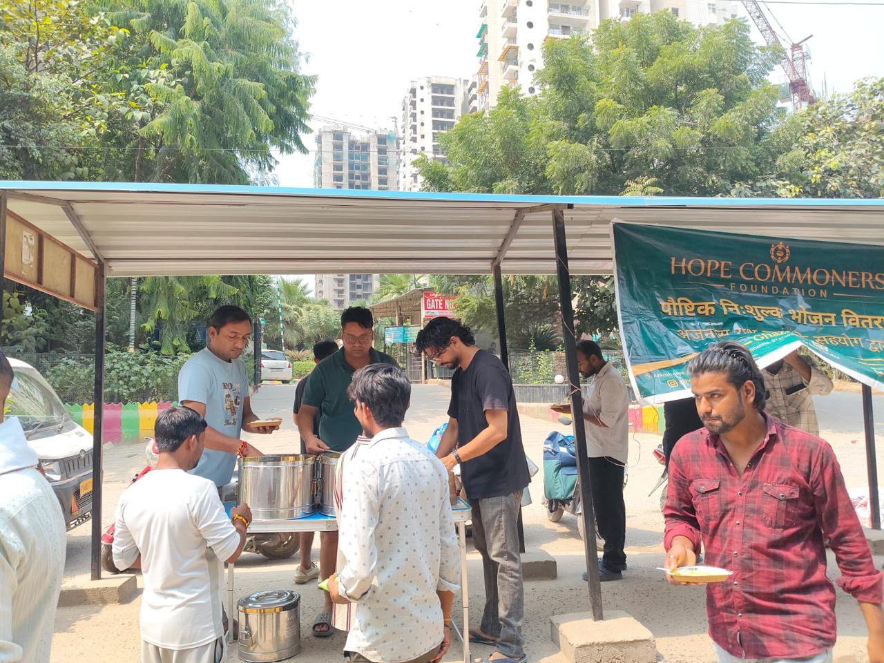 A man in a red checked shirt holds a plate as people serve from steel containers at a counter under a shelter.