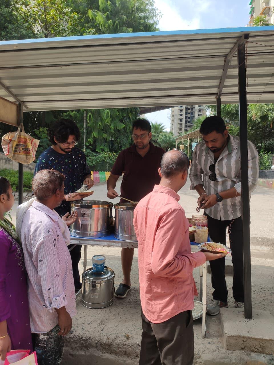 Three men serve from steel containers while a man in a pink shirt holds a plate and a woman in a patterned kurta stands at the left.