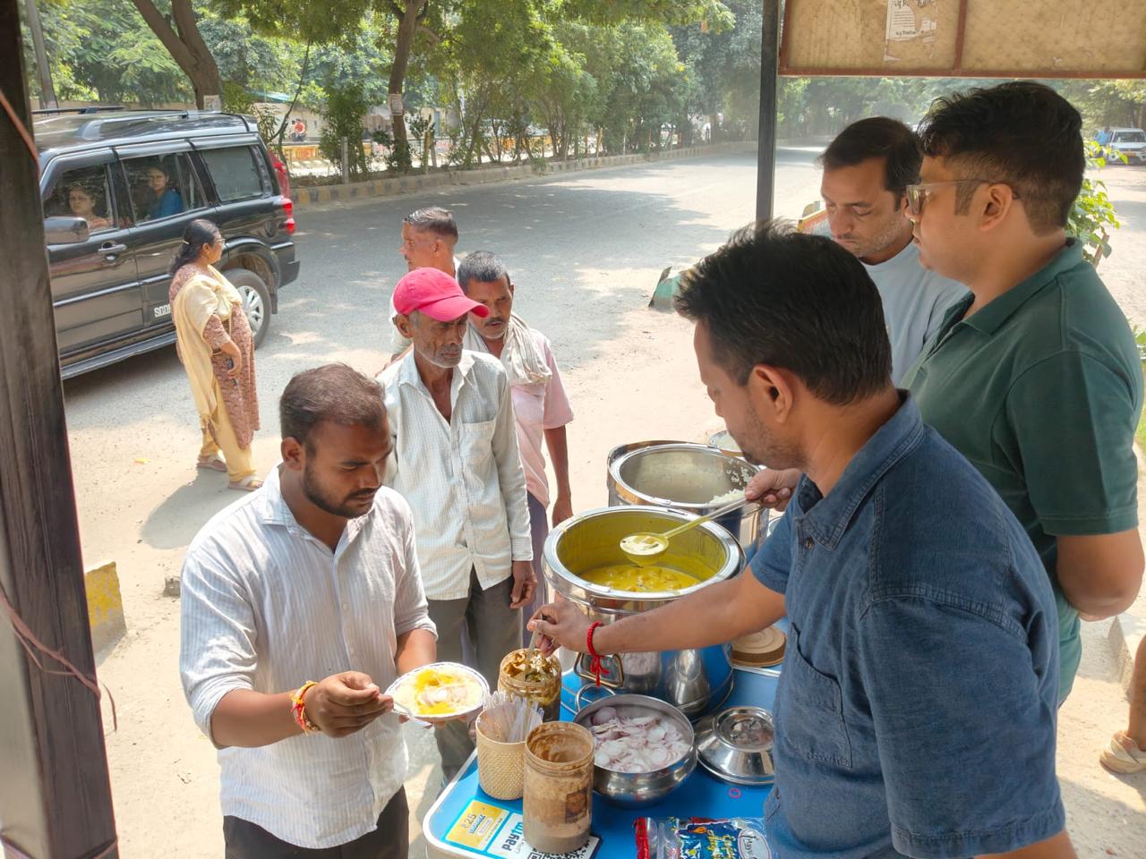 A man in a white shirt serves yellow dal from a steel pot at a counter while others wait, with a car and trees behind.