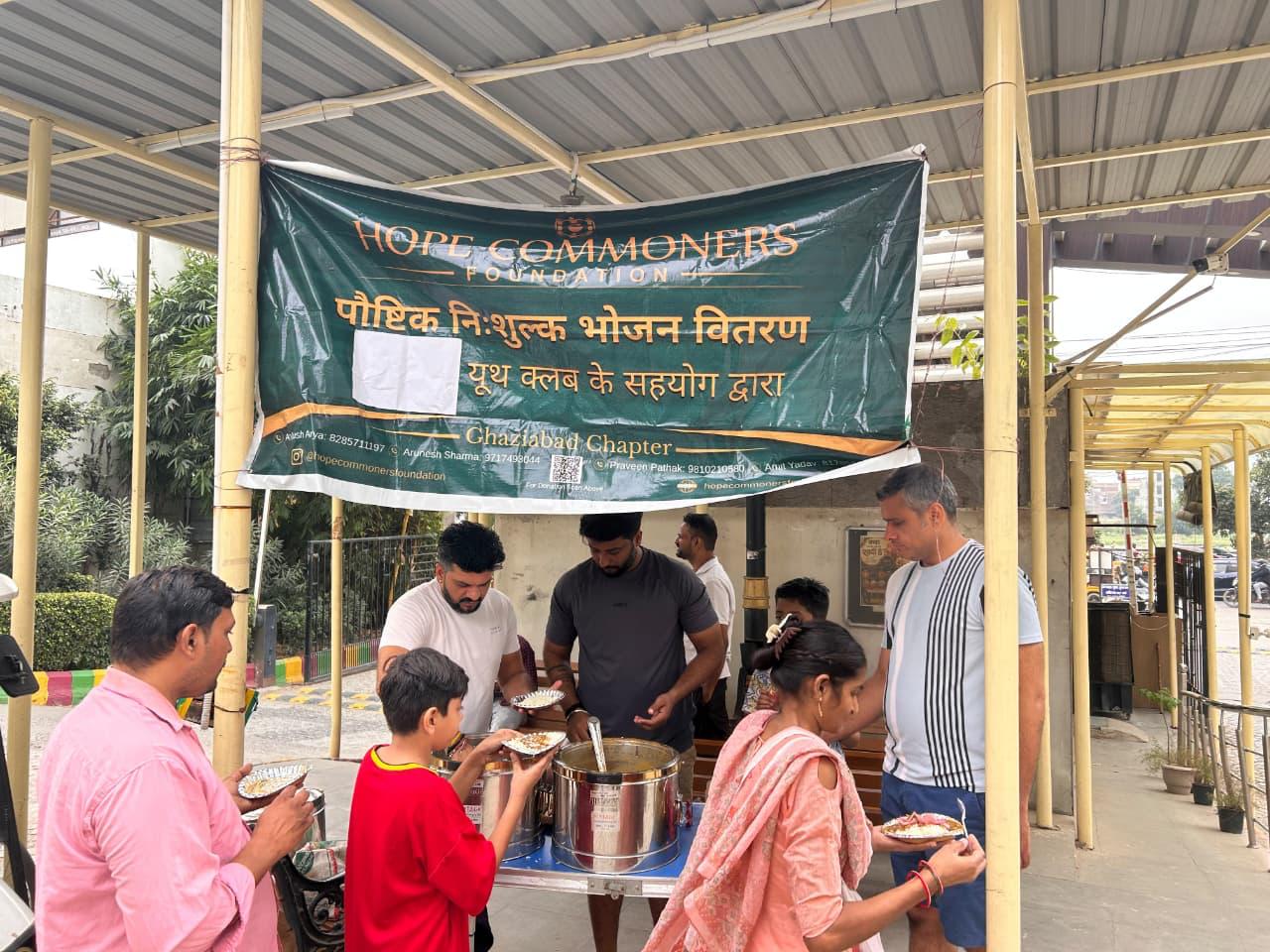A large green Hope Commoners Foundation banner hangs above a counter where people serve and children hold plates.