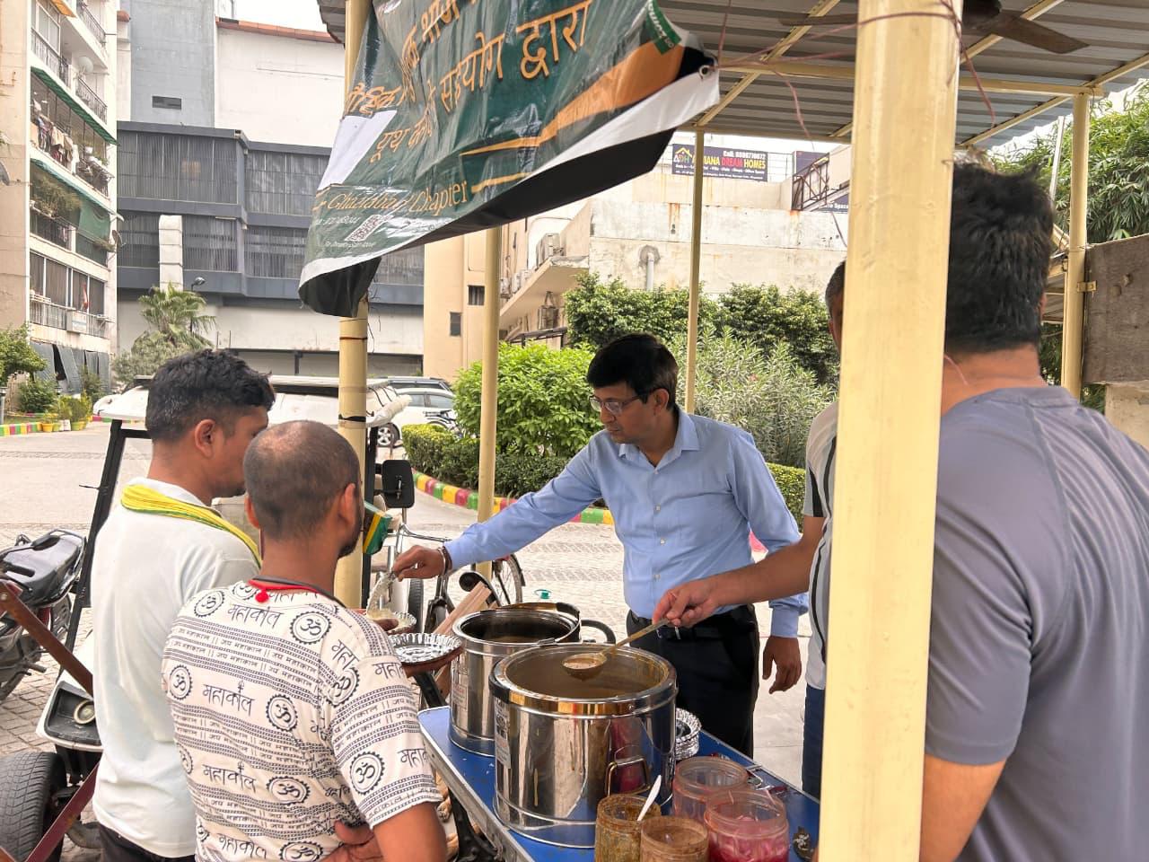 A man in a blue shirt serves from steel containers on a roadside counter, with a banner partly visible above and tall buildings beyond.
