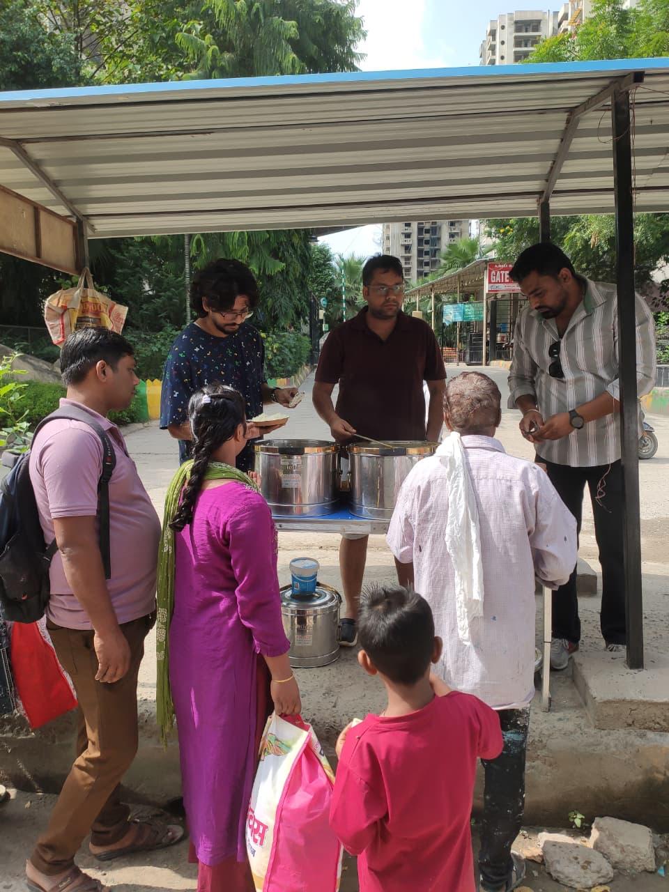 A woman in a purple kurta carrying a bag and a child in red stand at the counter while men serve from steel pots under a shelter.